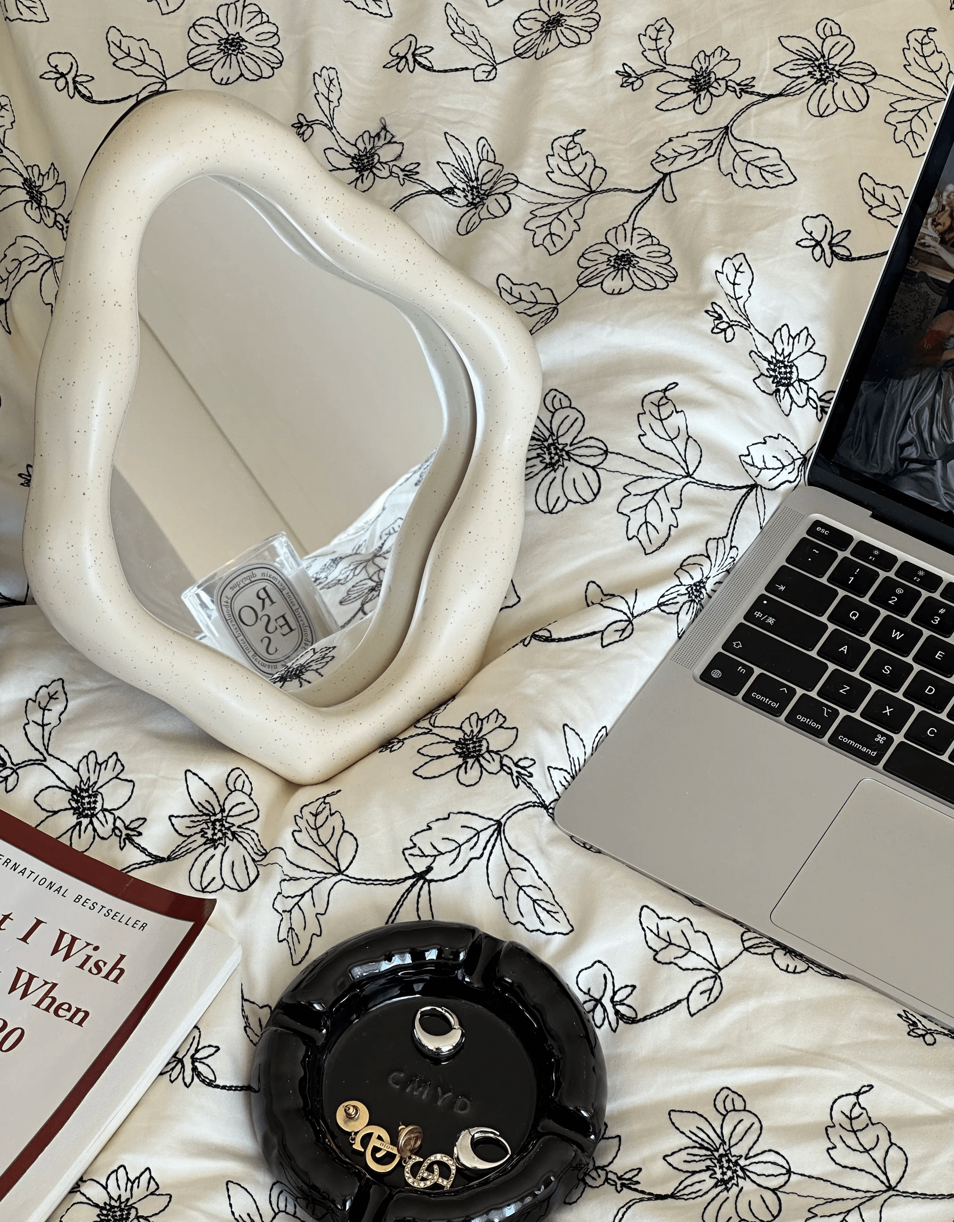 Elegant freestanding table mirror with curvy design on floral bedding beside a laptop and accessories.