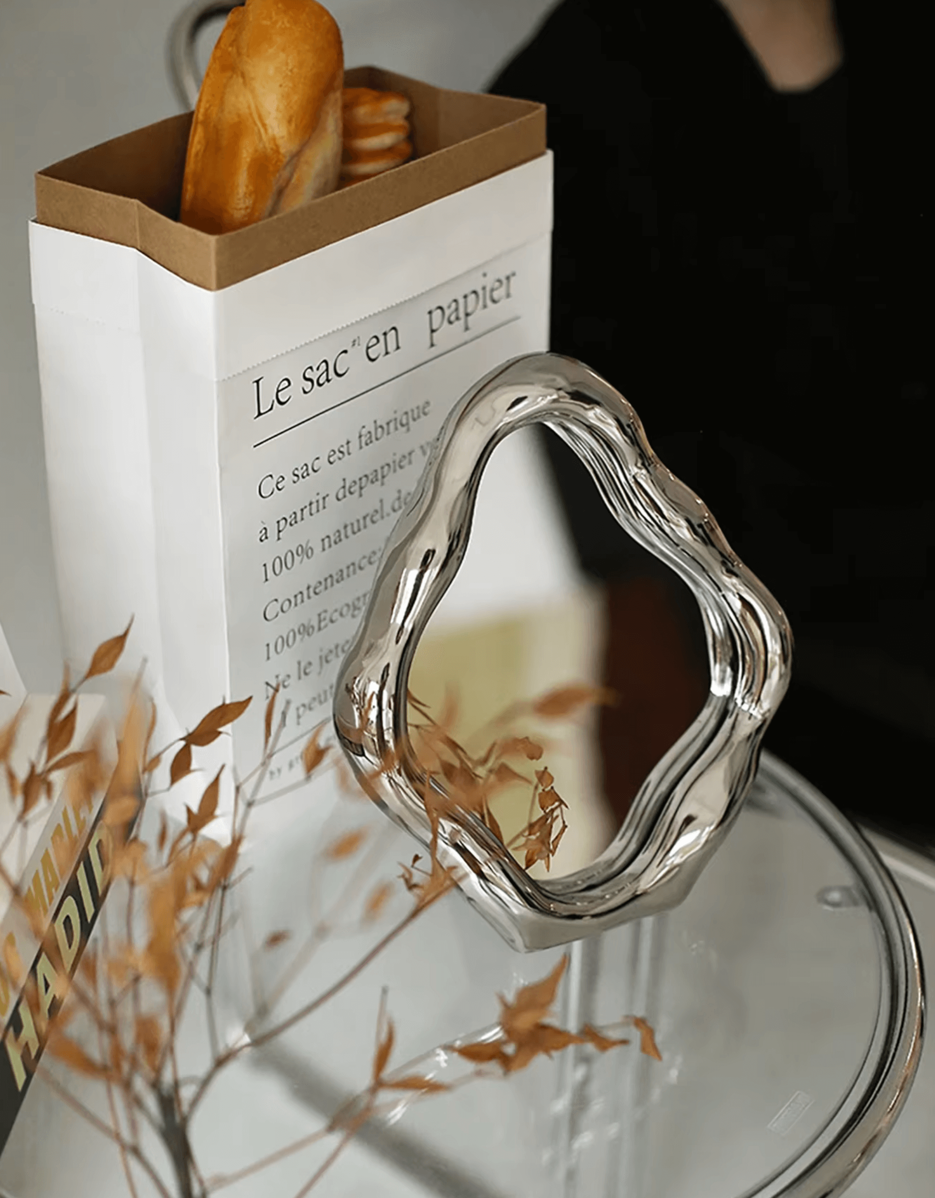 Decorative ceramic freestanding table mirror with elegant design next to a paper bag and dried plants on a glass table.