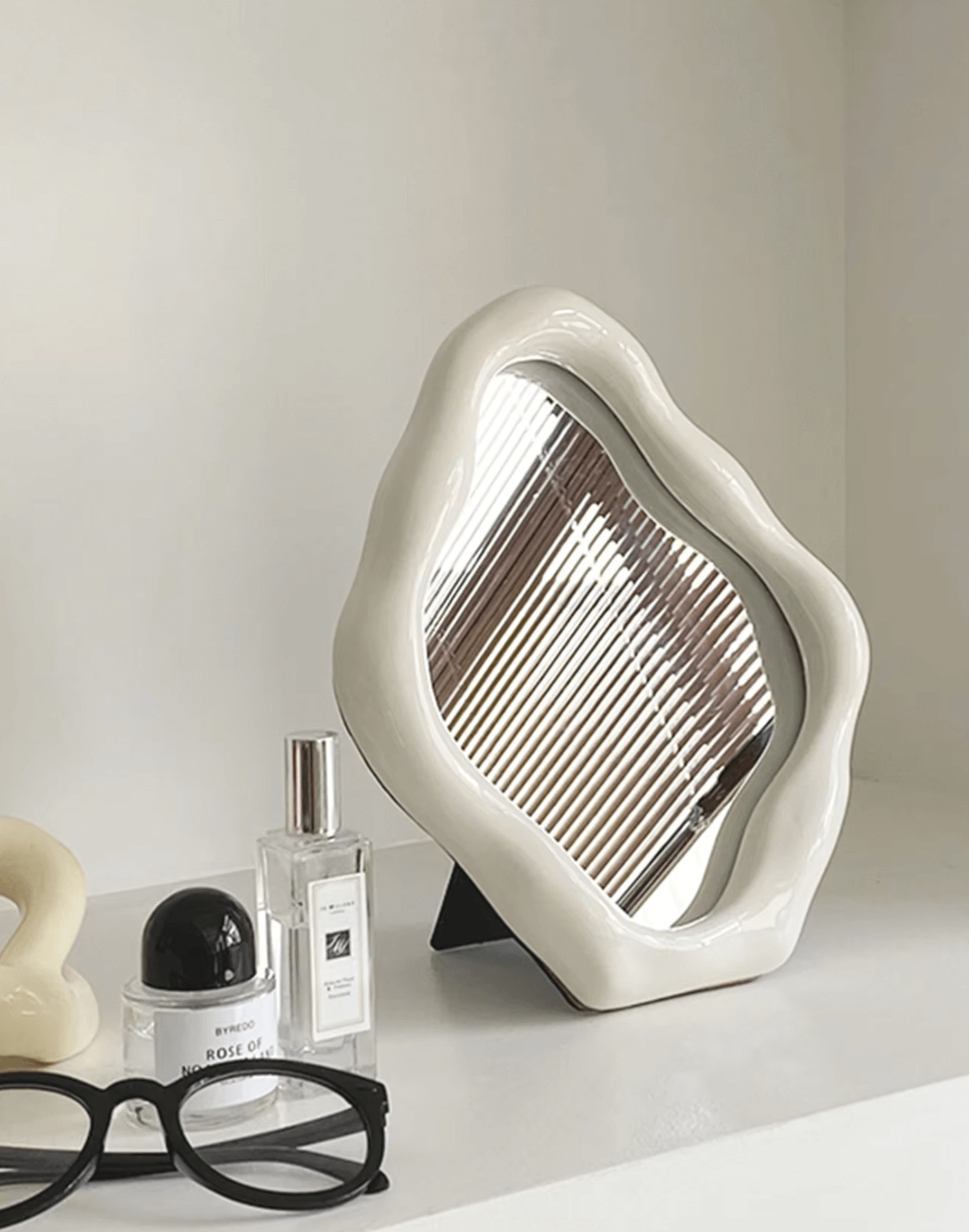 Elegant freestanding ceramic table mirror with unique design, surrounded by glasses and perfume bottle on a white shelf.