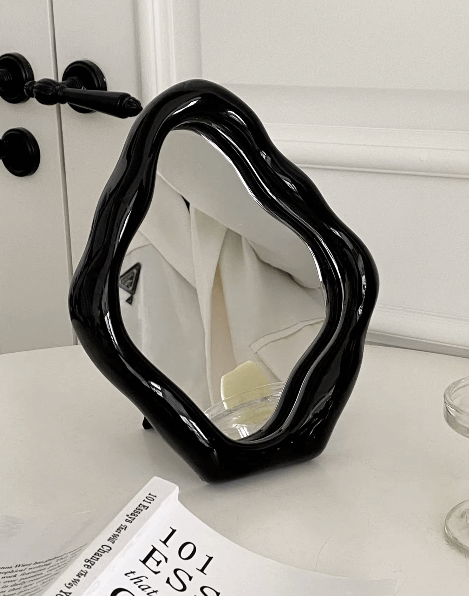 Elegant freestanding table mirror with artistic black ceramic frame on a white table near a door.