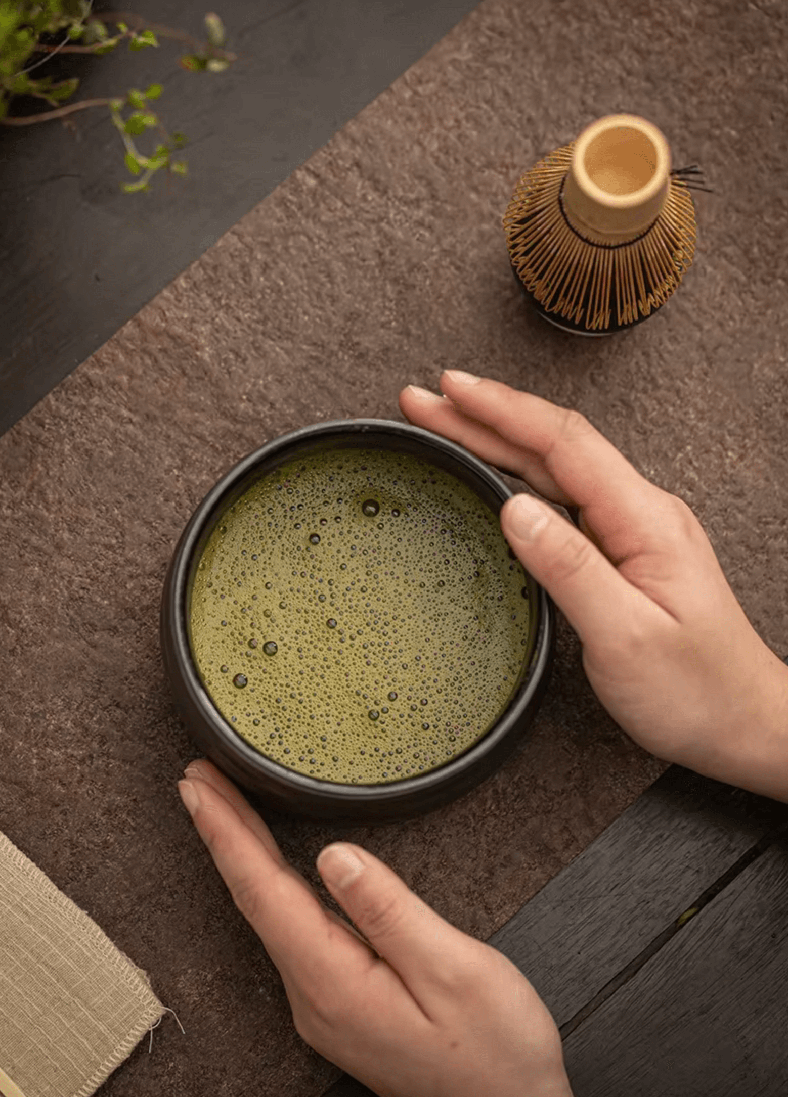 Hands holding a bowl of traditional matcha tea next to a bamboo whisk on a textured surface. Perfect for homemade tea ceremonies.