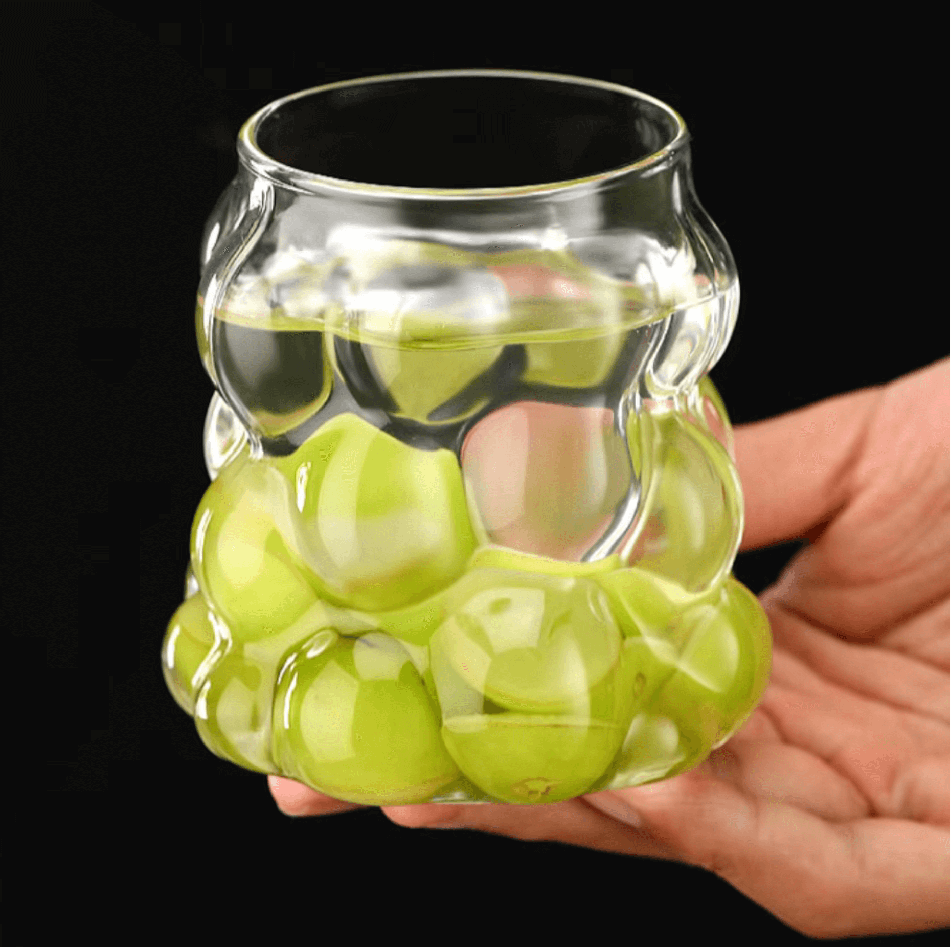 Hand holding a unique grape-shaped glass cup filled with green grapes and water on a black background.