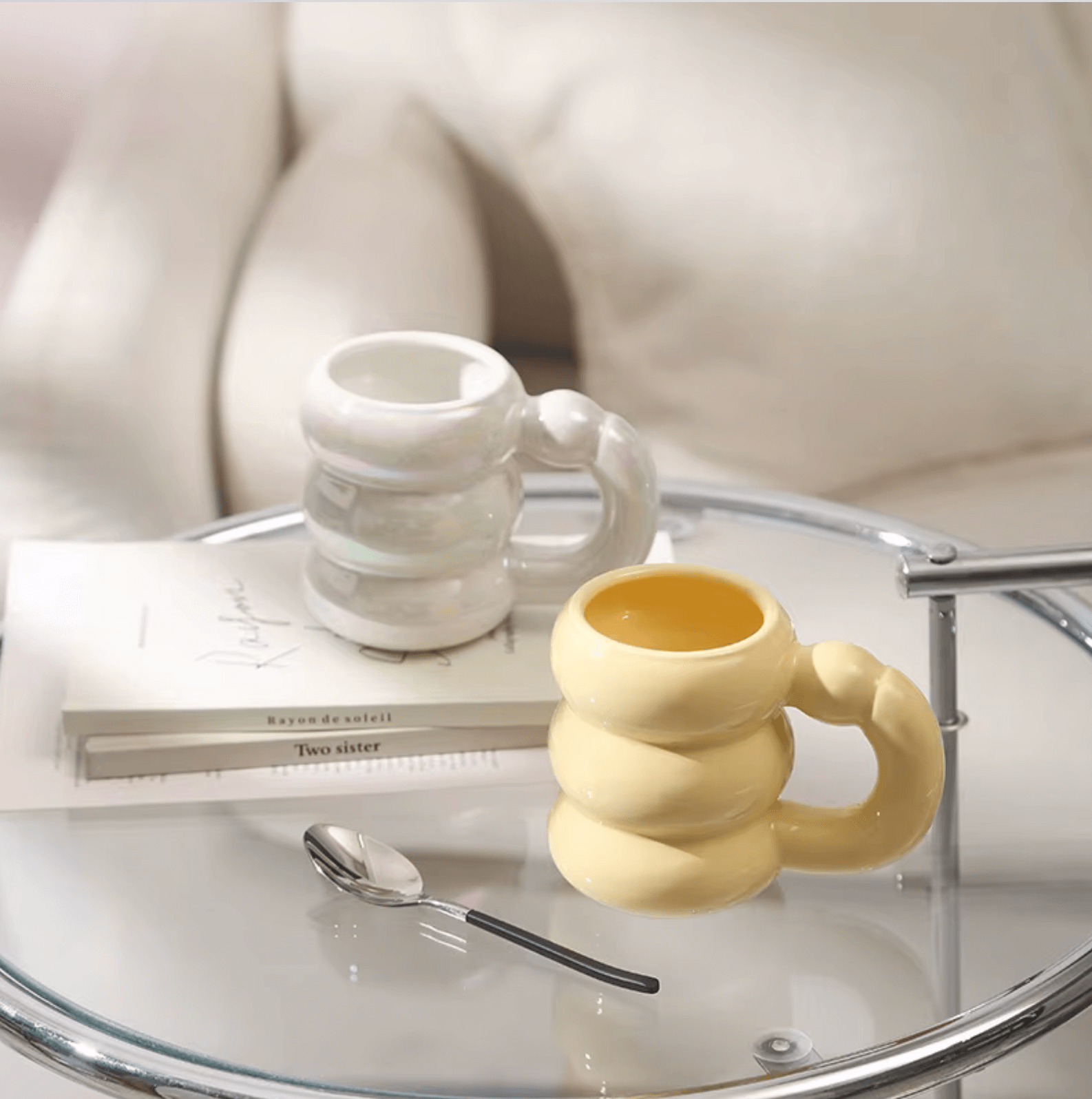 Two stylish mugs in modern Korean design on a glass table next to books and a spoon, perfect for coffee or tea enthusiasts.