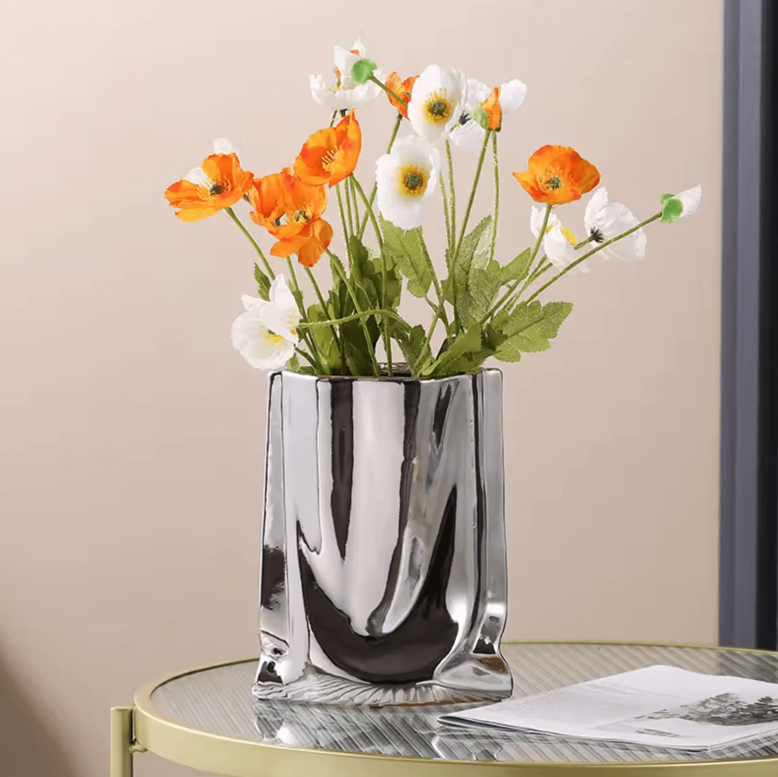 Silberne Blumenvase aus Keramik mit glänzendem Finish, gefüllt mit orange und weißen Blumen, auf einem runden Tisch