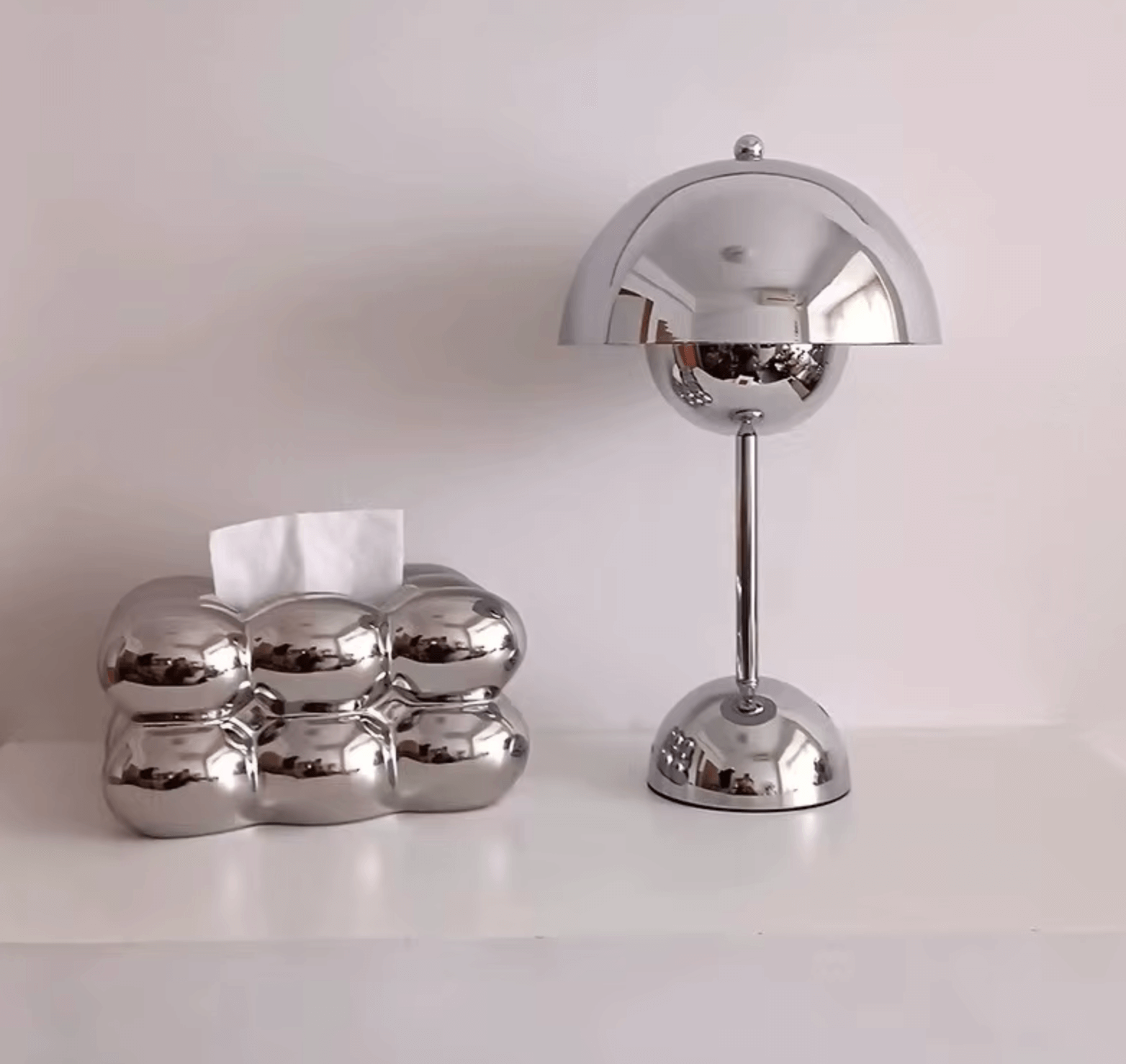 Dänische Blumenlampe aus Edelstahl neben verspiegeltem Tissue-Box-Halter auf einem weißen Regal.