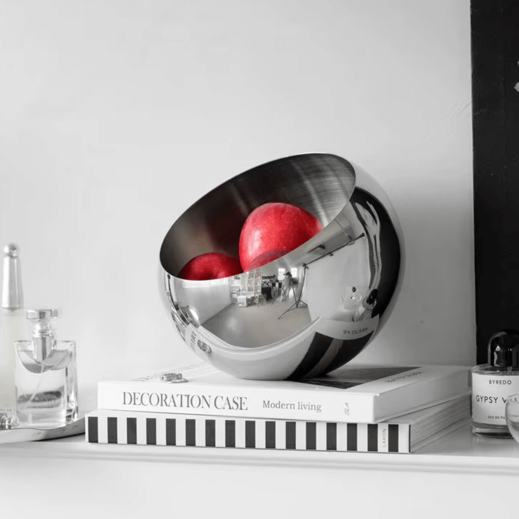 Modern stainless steel bowl with red apples, minimalistic design, placed on a shelf with books and decorative items.