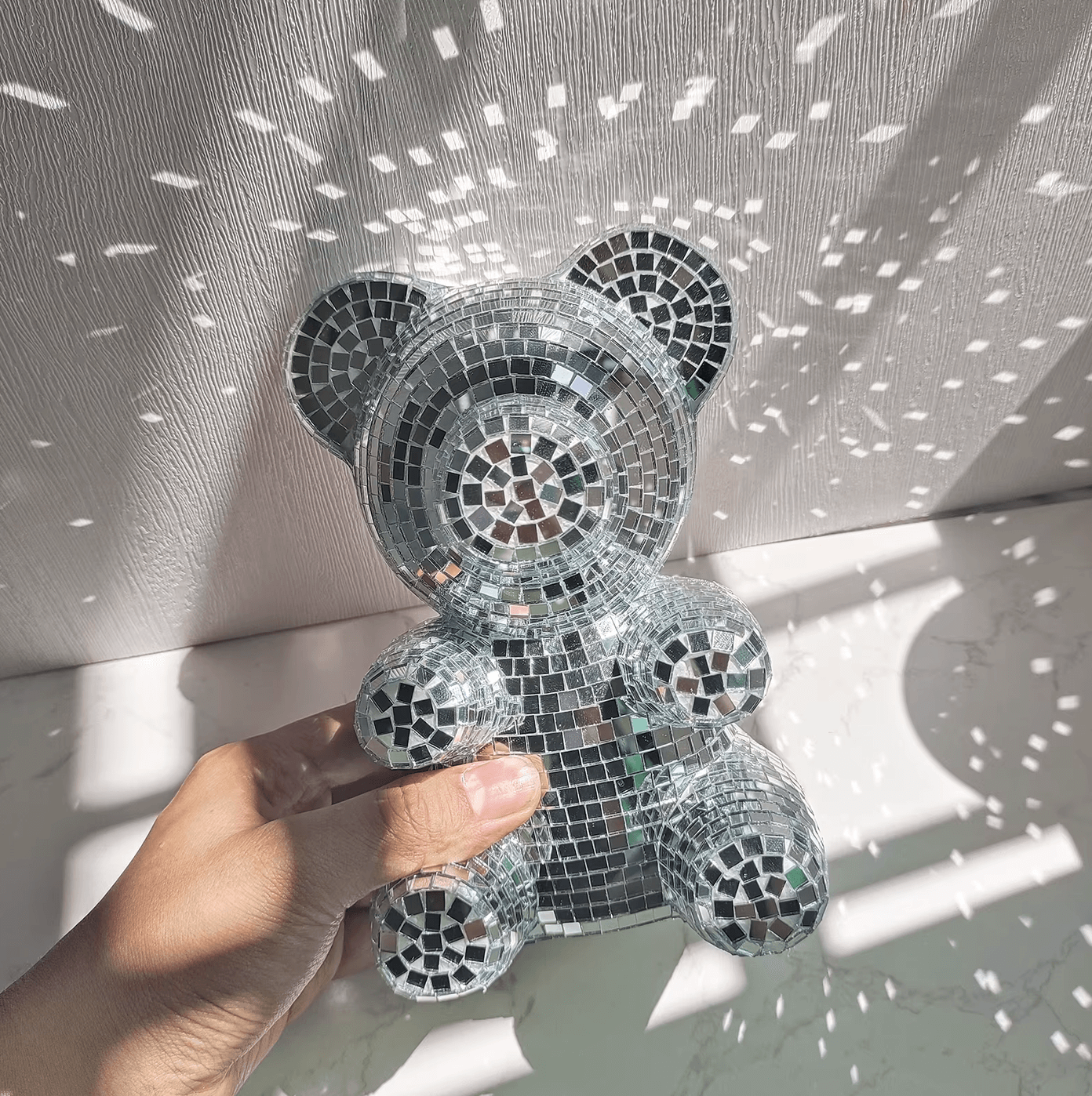 Hand holding Disco-Bär decoration with mirror tiles reflecting light, blending cute bear design and disco charm, retro style interior decor.