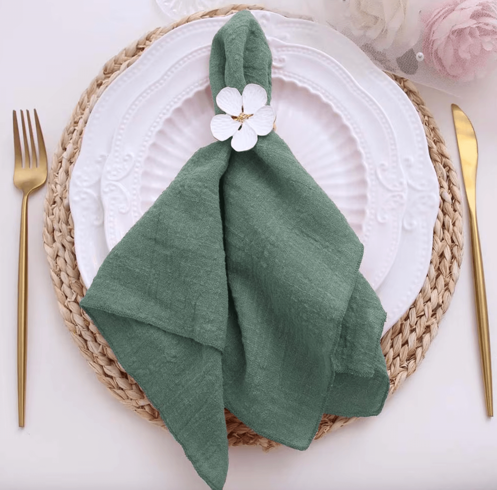 Elegant green cotton napkin with flower decoration on a white plate, set on a woven placemat with gold cutlery for a stylish table setting.
