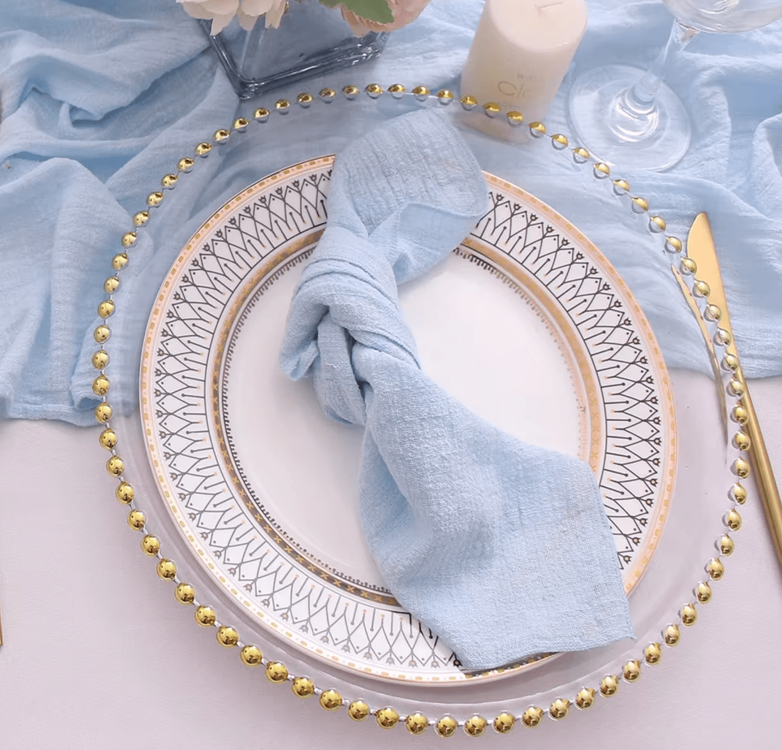 Elegant table setting with a blue cotton napkin on a decorative plate, surrounded by gold cutlery and a glass.