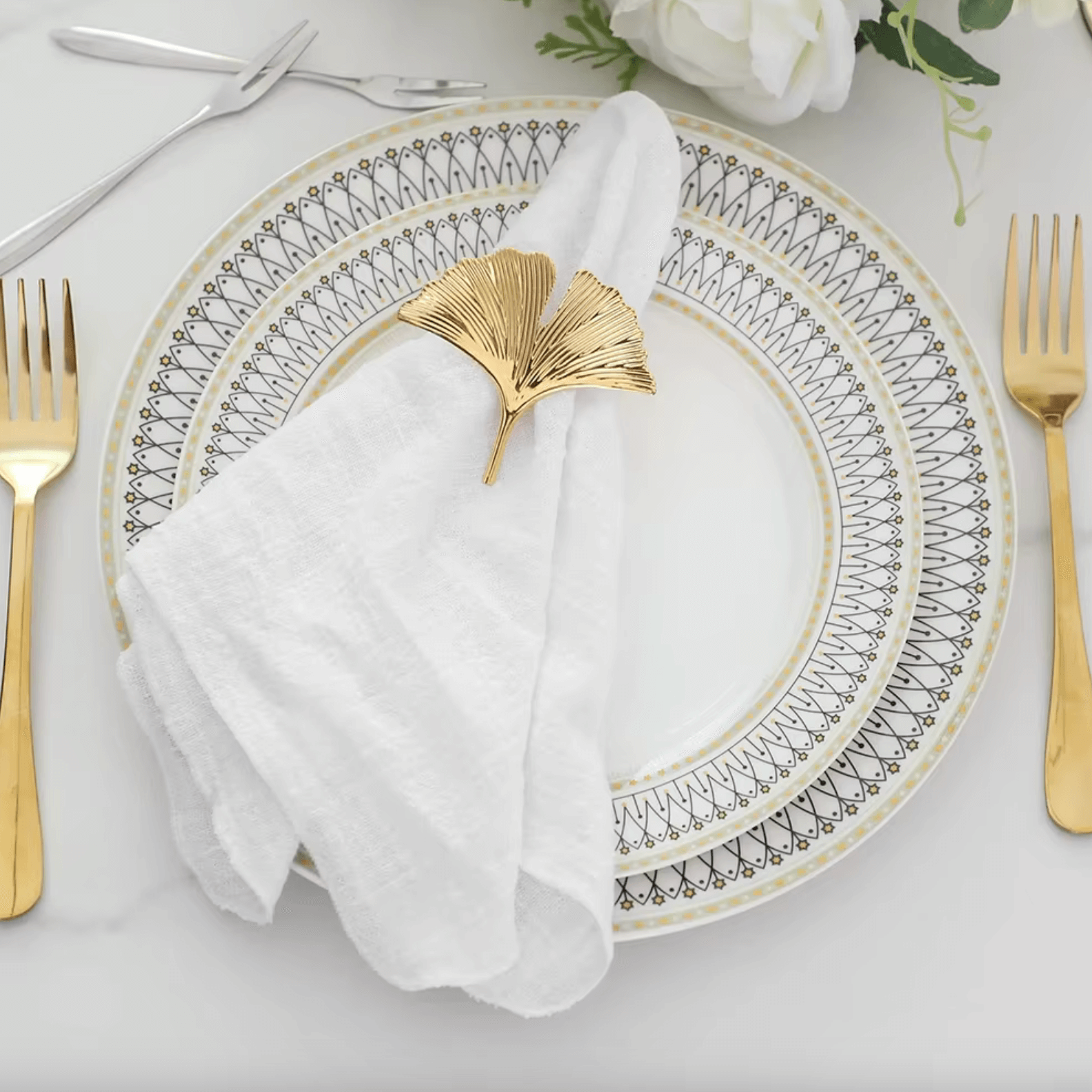Elegant cotton napkin with gold leaf holder on decorative plate set, surrounded by luxurious cutlery and floral decor.