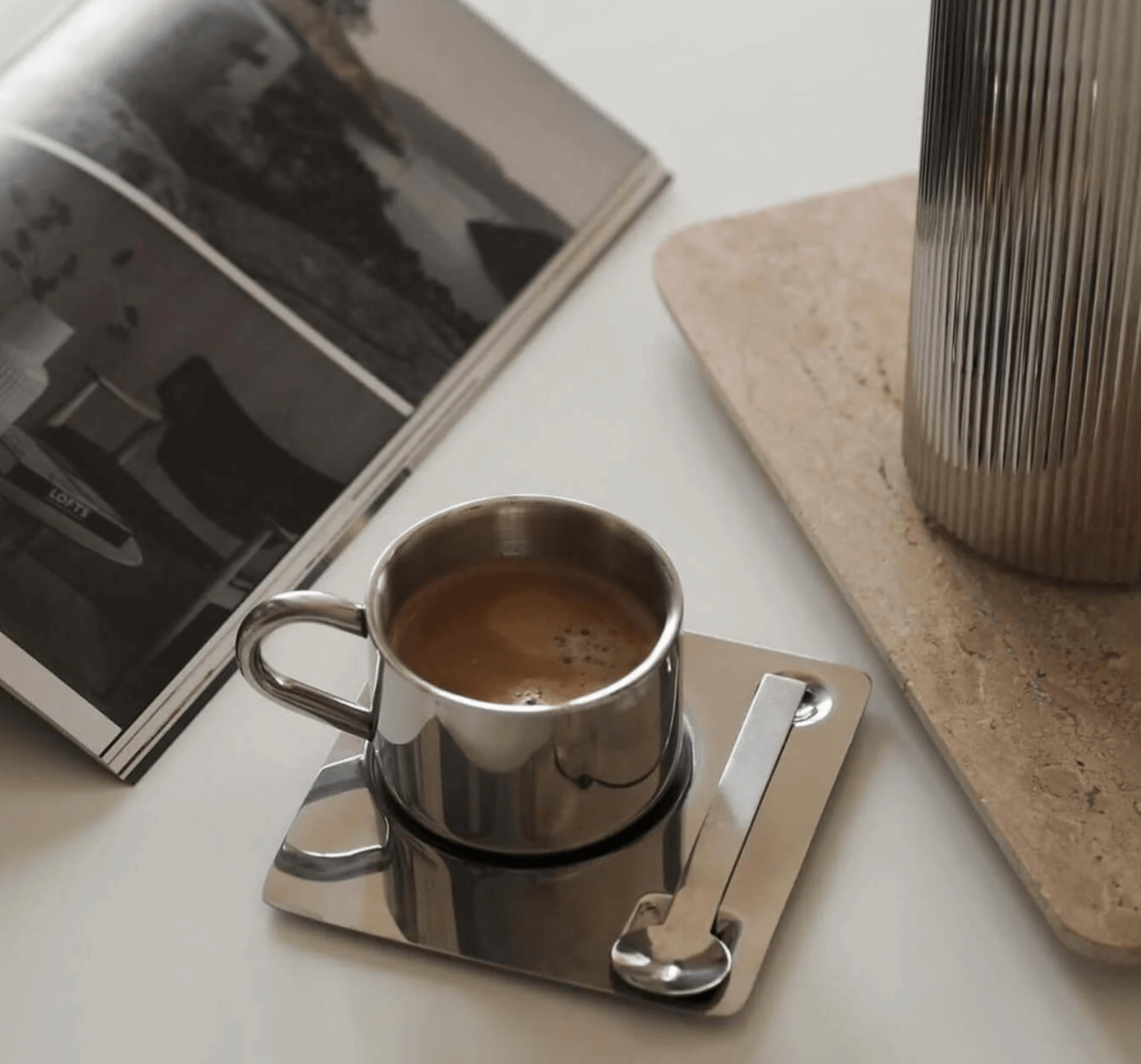 Edelstahl-Kaffeeset mit doppelwandiger Tasse und Untertasse, luxuriös und eckig, auf einem Tisch neben einer Zeitschrift.