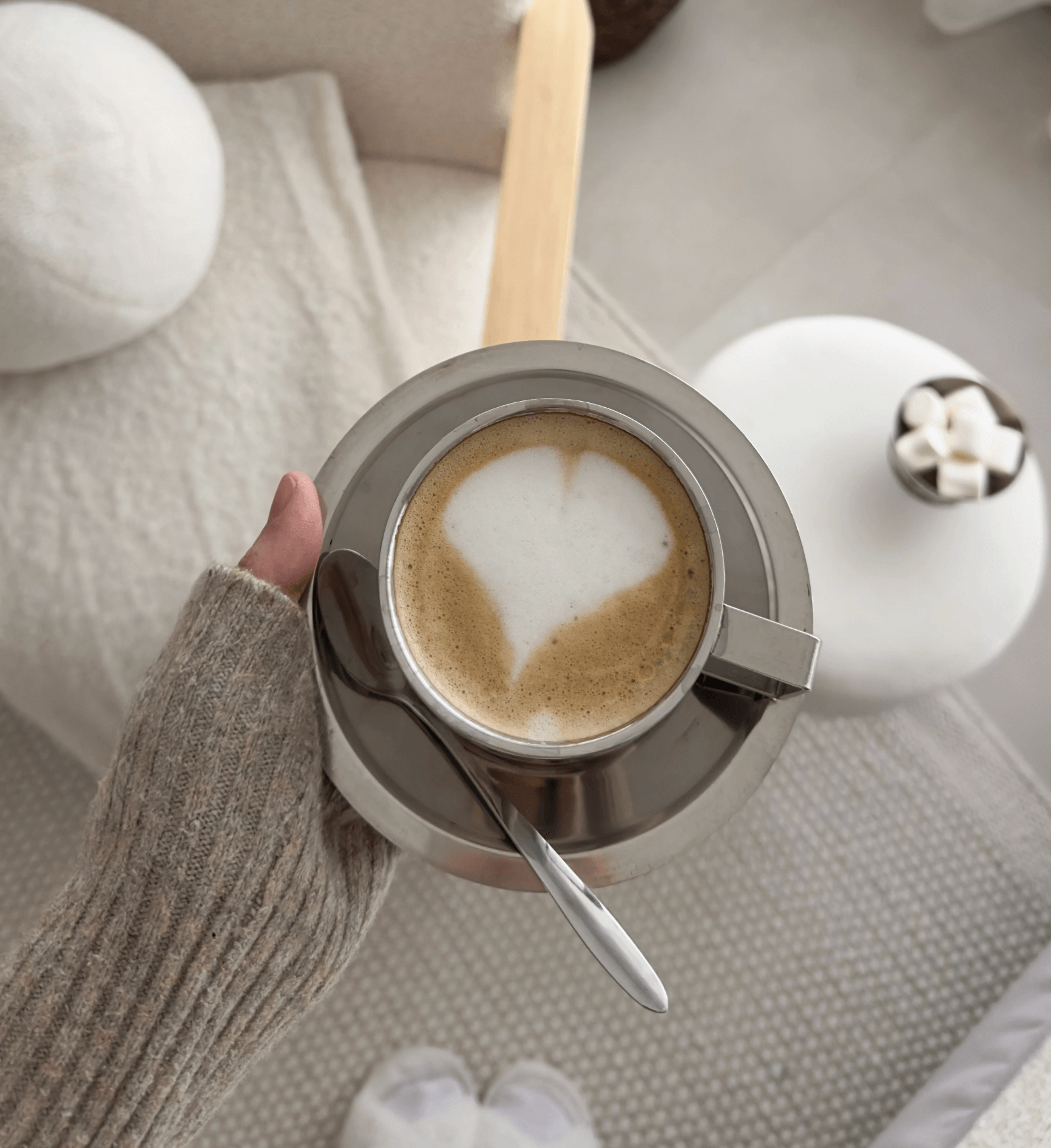 Person holding a cup of coffee with heart-shaped foam in a minimalist stainless steel set.