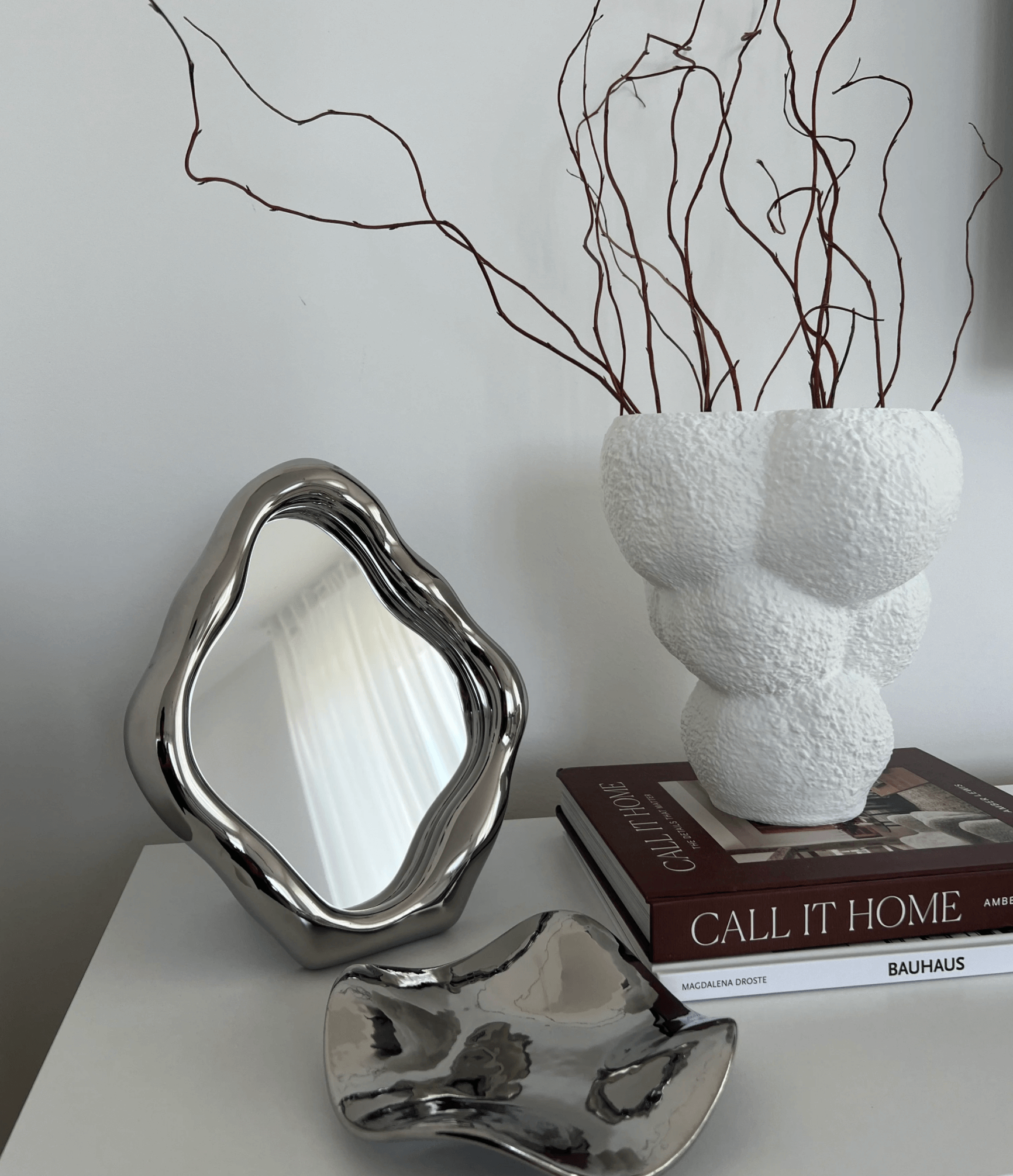 Elegant freestanding table mirror on a white surface next to sculptural vase and design books, showcasing stylish interior decor arrangement.