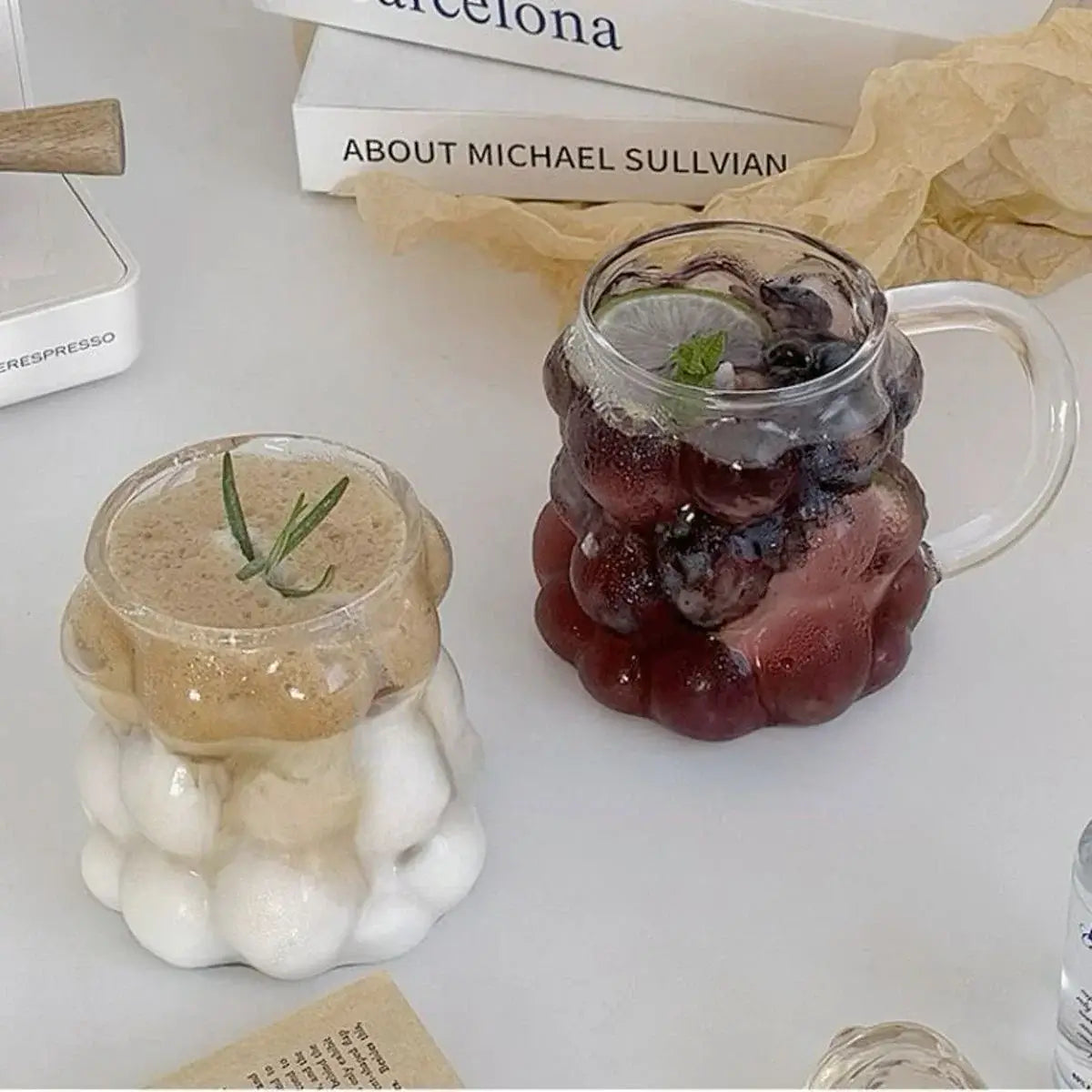 Two bubble glass mugs with drinks; one with a creamy beverage and rosemary, the other with a cold berry drink and mint garnish.