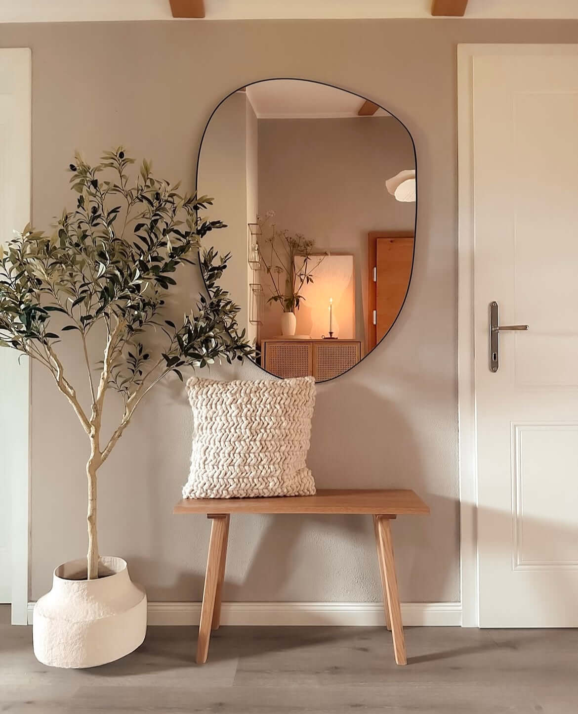 Modern interior with artificial olive tree in a white pot beside a bench with a textured cushion, reflecting in an organic-shaped mirror.