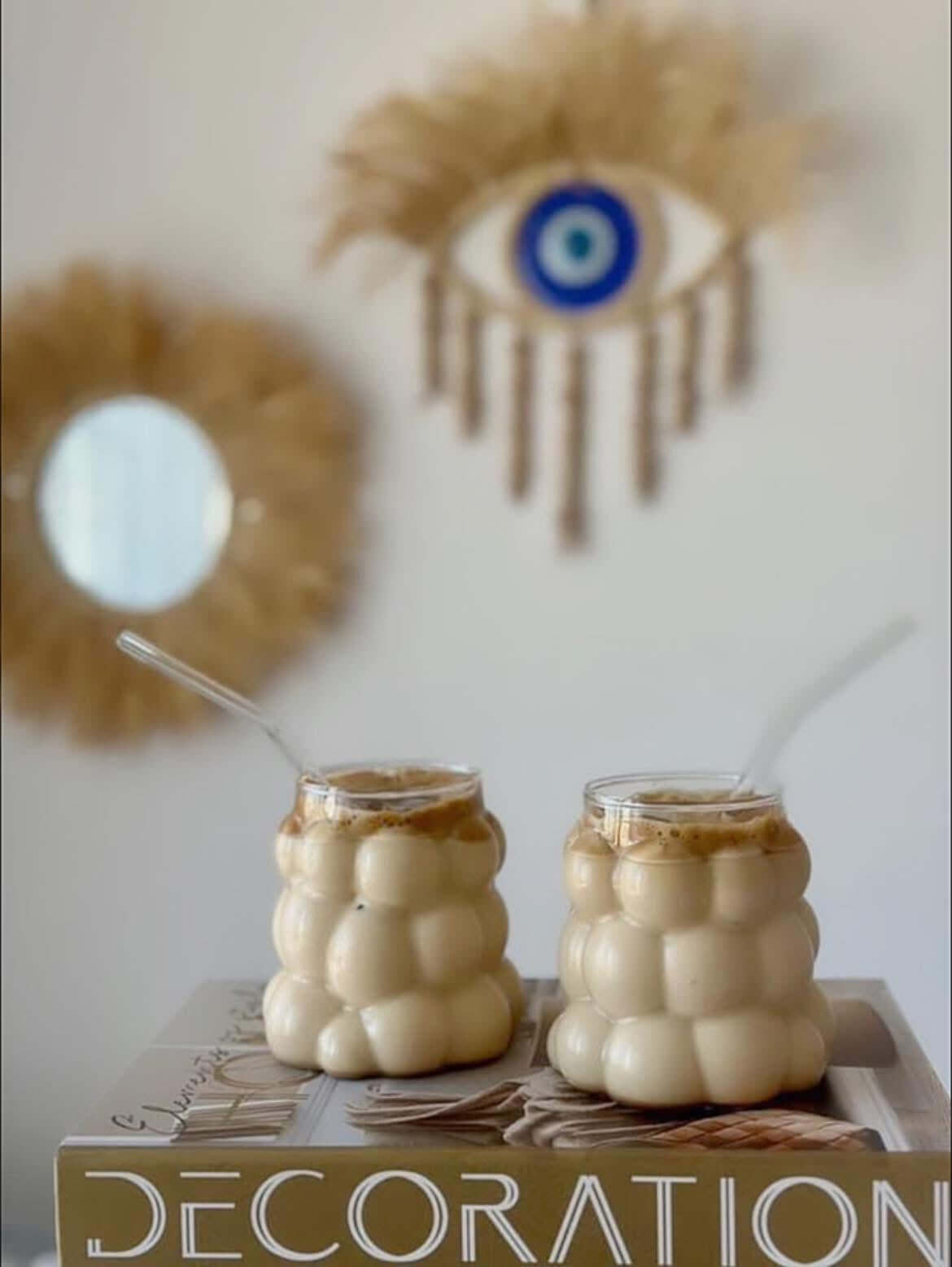 Two bubble glasses filled with iced coffee on a book, with eye-shaped wall art in the background.