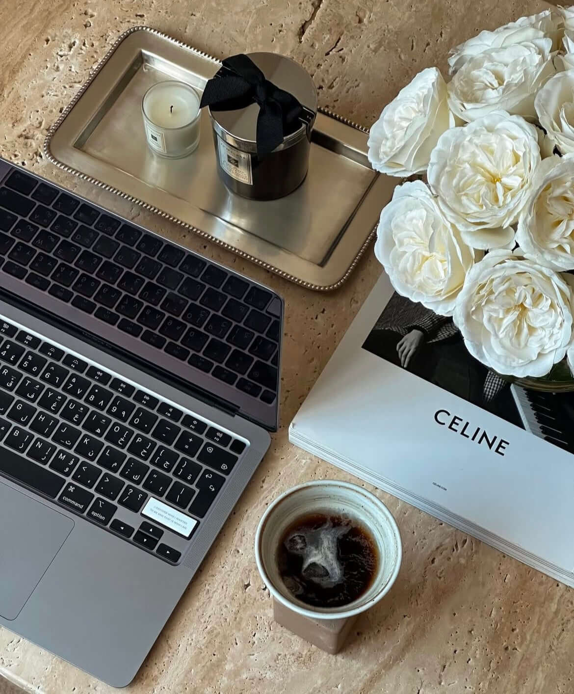 Stylish workspace with laptop, coffee, Celine book, scented candles, and white roses on a marble surface.
