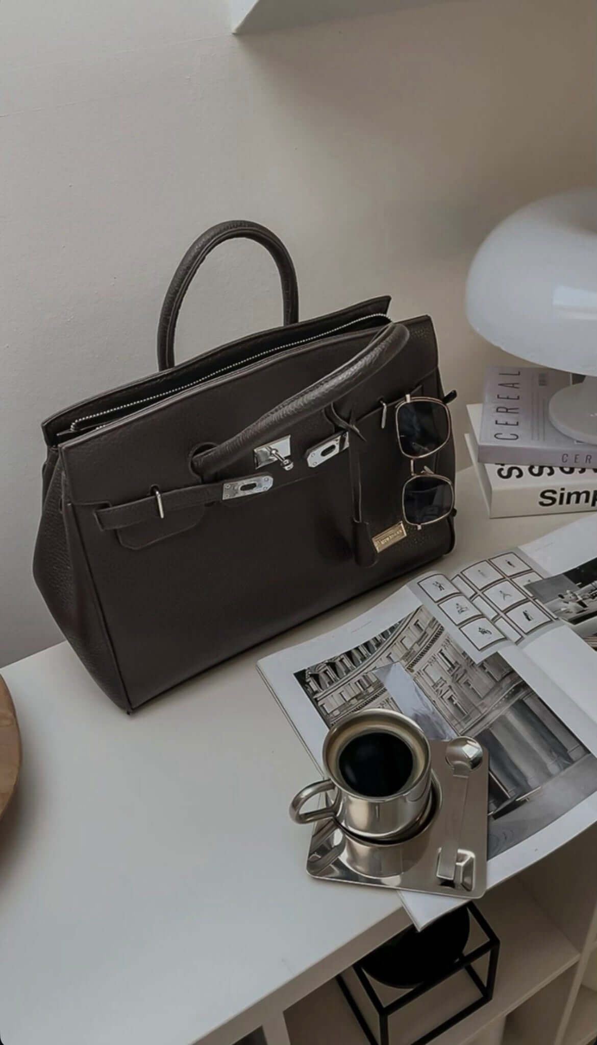 Stylish gray handbag with sunglasses and magazines beside a luxury stainless steel coffee set on a table.
