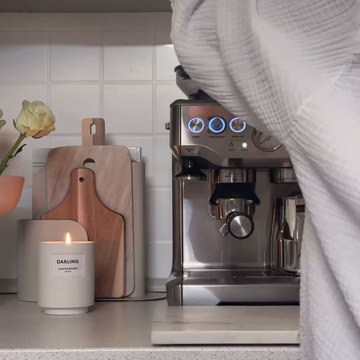 Person using a stainless steel espresso machine on a kitchen counter with cutting boards, a candle, and a flower vase.