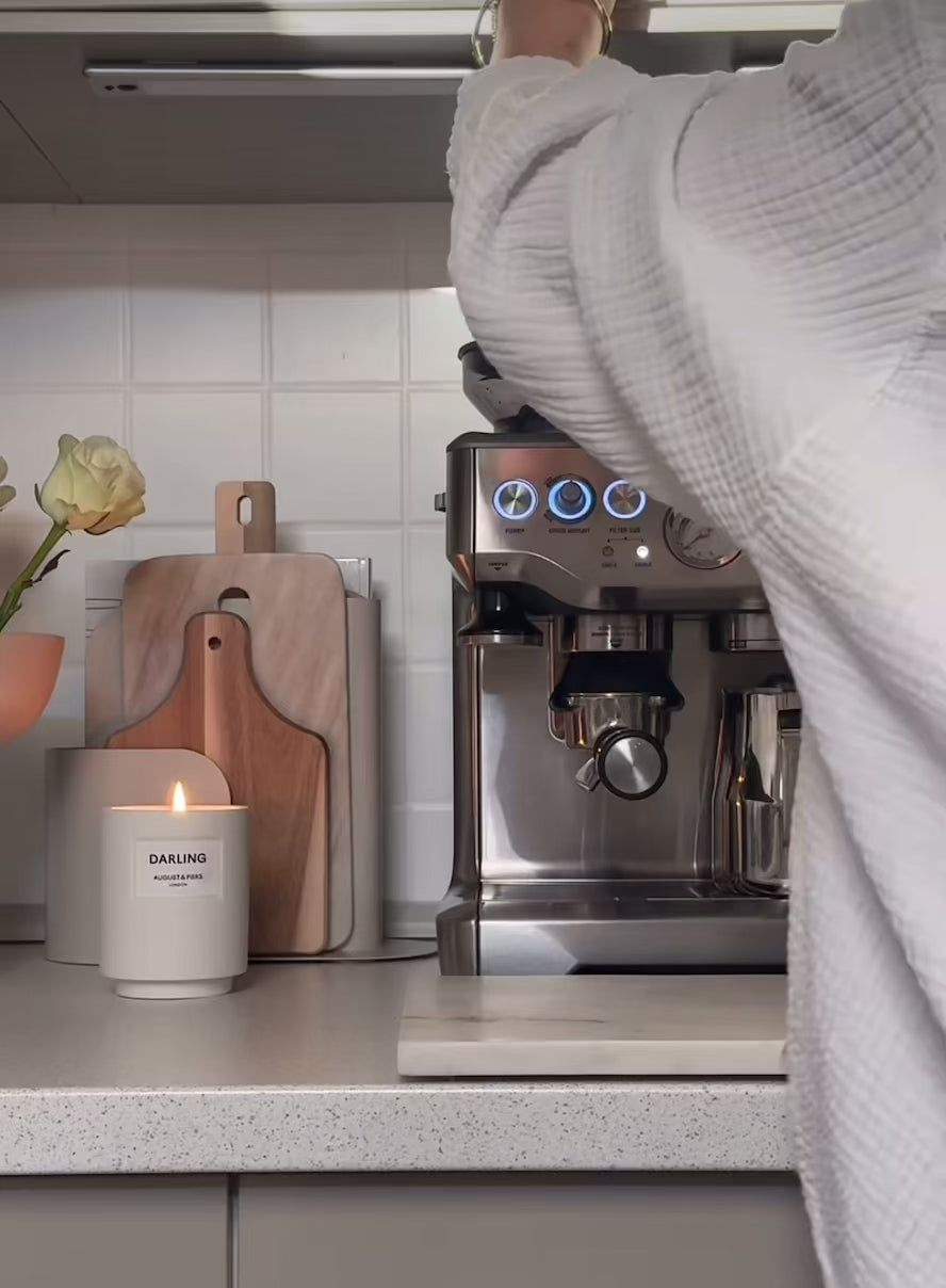 Person using a stainless steel espresso machine on a kitchen counter with cutting boards, a candle, and a flower vase.