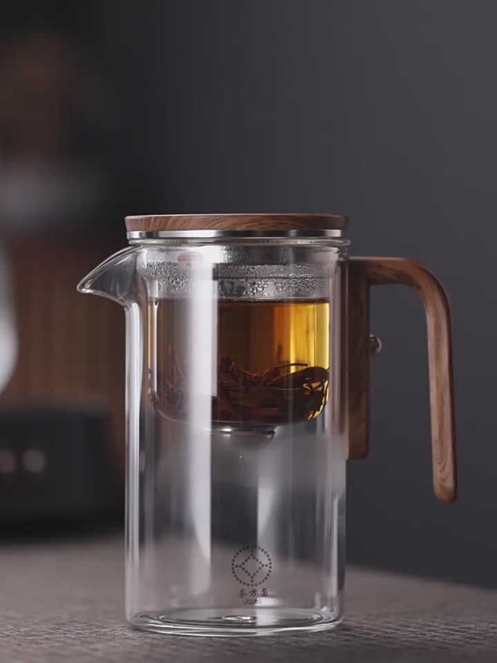 Teekanne "Zen Brew" aus Glas mit Holzelement, ideal für heißen und kalten Tee, steht auf einem Tisch vor dunklem Hintergrund.